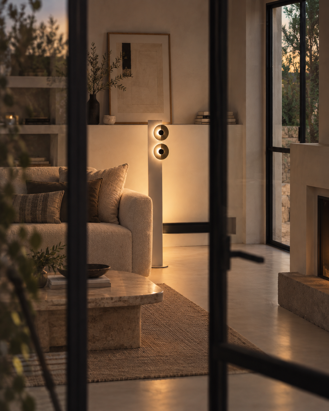A tall, minimalist floor-standing speaker with two circular driver grilles, glowing with warm light in a modern, softly lit living room. The room features a beige sofa, a stone coffee table, and a fireplace in the background, viewed through dark window panes.