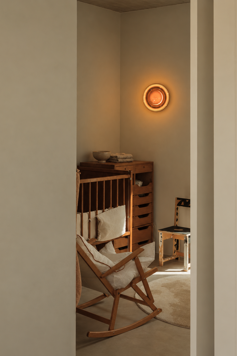 A warm, minimalist nursery corner viewed through a doorway, featuring a wooden rocking chair with a white cushion, a wooden changing table, and a glowing circular wall light on a beige wall.