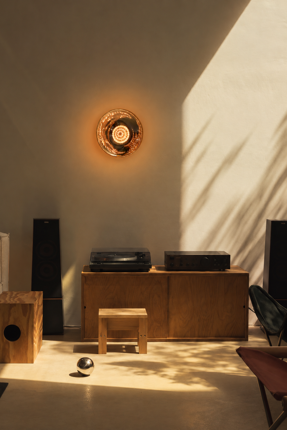 A warm, modern interior scene with a circular wall light glowing on a beige wall, sunlight casting leaf-like shadows, and wooden furniture including a cabinet, stool, speakers, and a small wooden box.