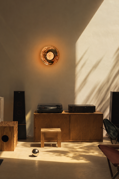 A warm, modern interior scene with a circular wall light glowing on a beige wall, sunlight casting leaf-like shadows, and wooden furniture including a cabinet, stool, speakers, and a small wooden box.