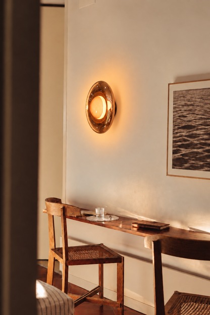 A warm, oval-shaped wall sconce with a metallic gold finish and glowing light, mounted on a cream-colored wall in a cozy, warmly lit room with a wooden desk and chair.
