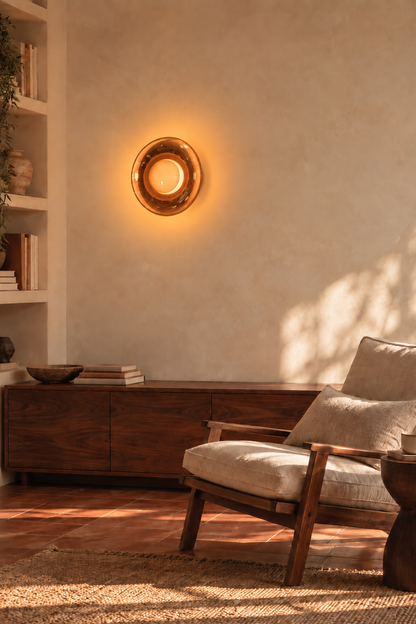 A warm, circular wall sconce with a textured bronze finish and glowing candle-like light, mounted on a textured beige plaster wall in a beautifully styled room with a wooden console, a mid-century modern armchair, and warm sunlight casting shadows.