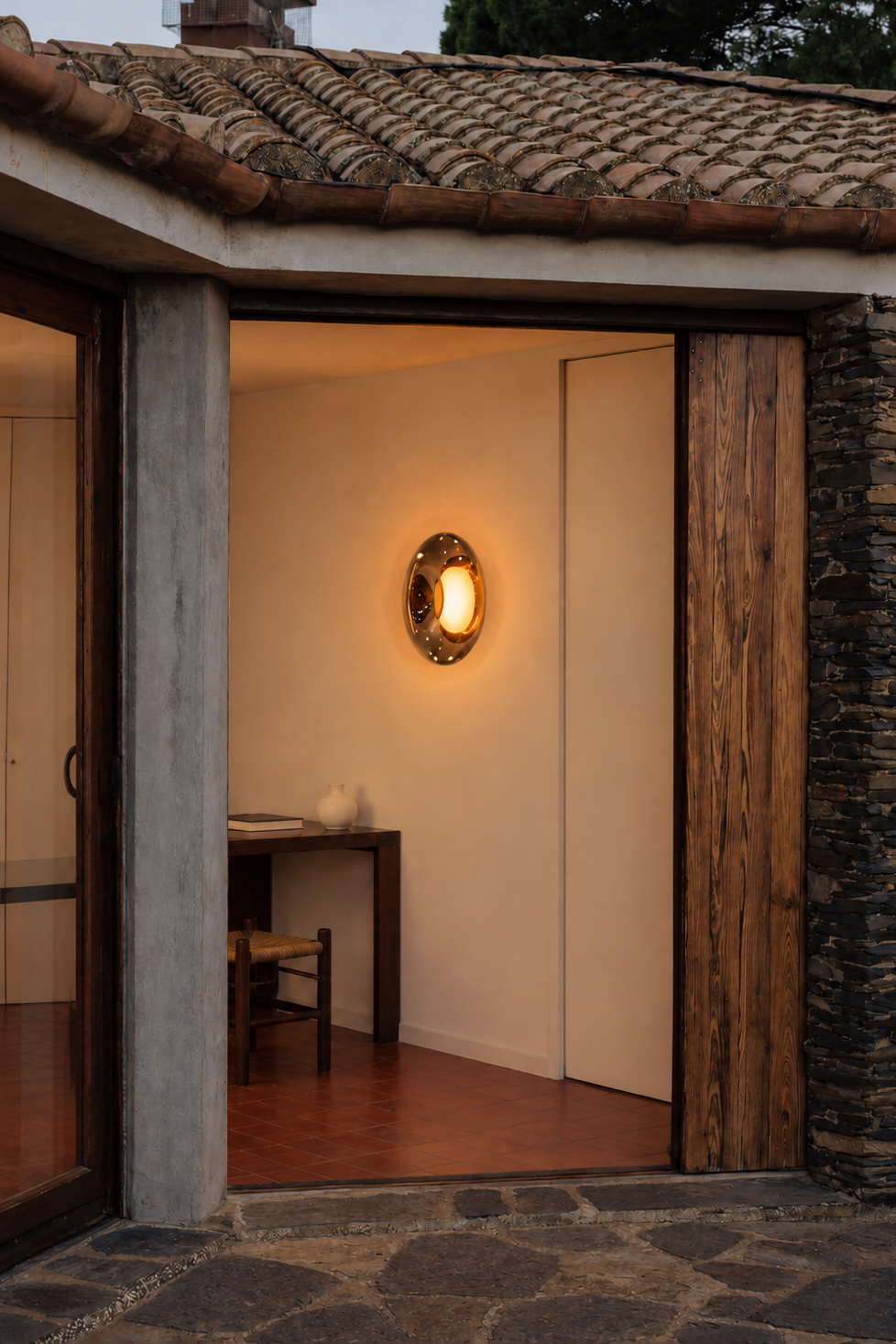 A warm, oval wall sconce with a gold-toned frame and glowing light, mounted on an off-white wall inside a rustic home entryway with wooden doors and a tiled roof.