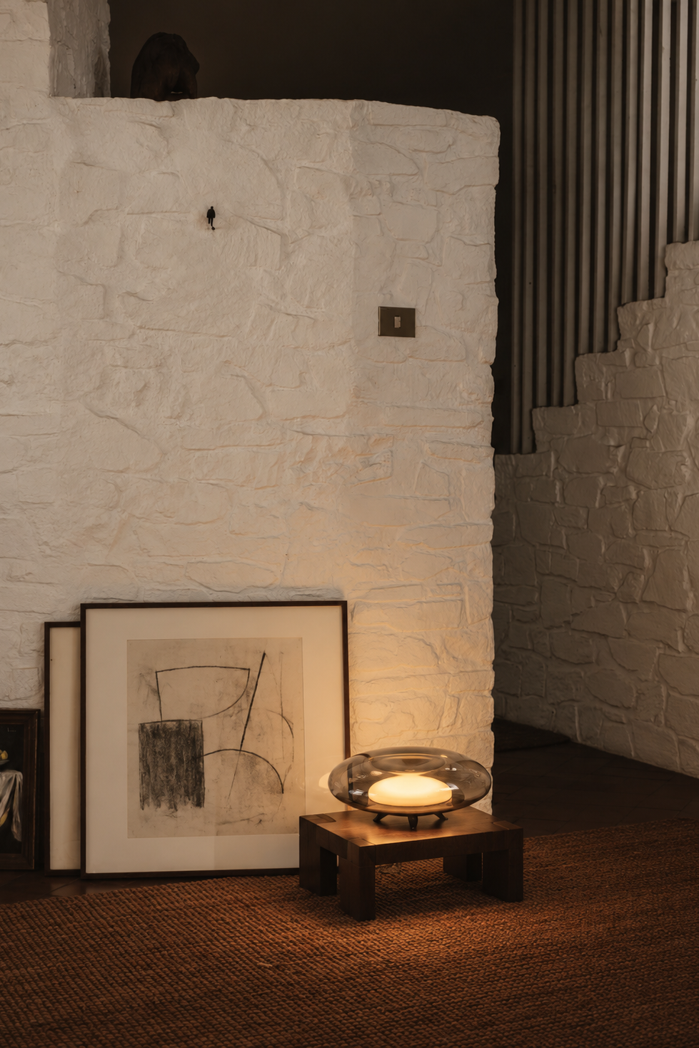 A warm, moody interior scene featuring a white textured stone wall, a small wooden table holding a glowing glass bowl candle, and two framed black-and-white sketches leaning against the wall on a brown woven rug.