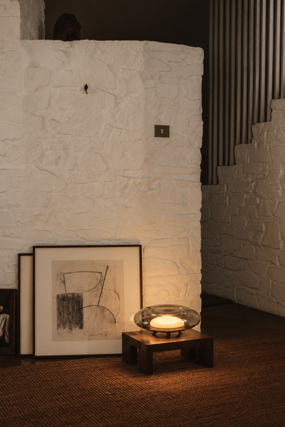 A warm, moody interior scene featuring a white textured stone wall, a small wooden table holding a glowing glass bowl candle, and two framed black-and-white sketches leaning against the wall on a brown woven rug.