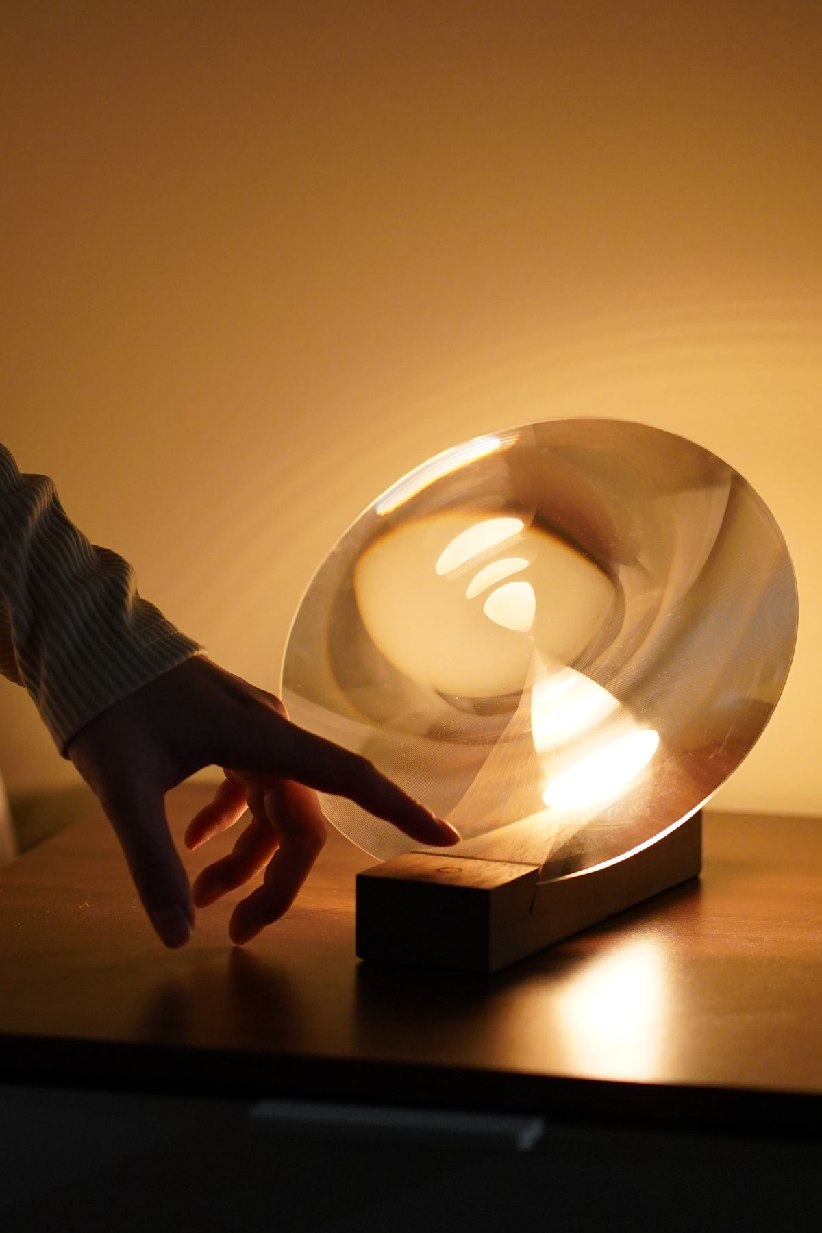 Hand touching a modern, round light fixture on a wooden surface with a warm glow.