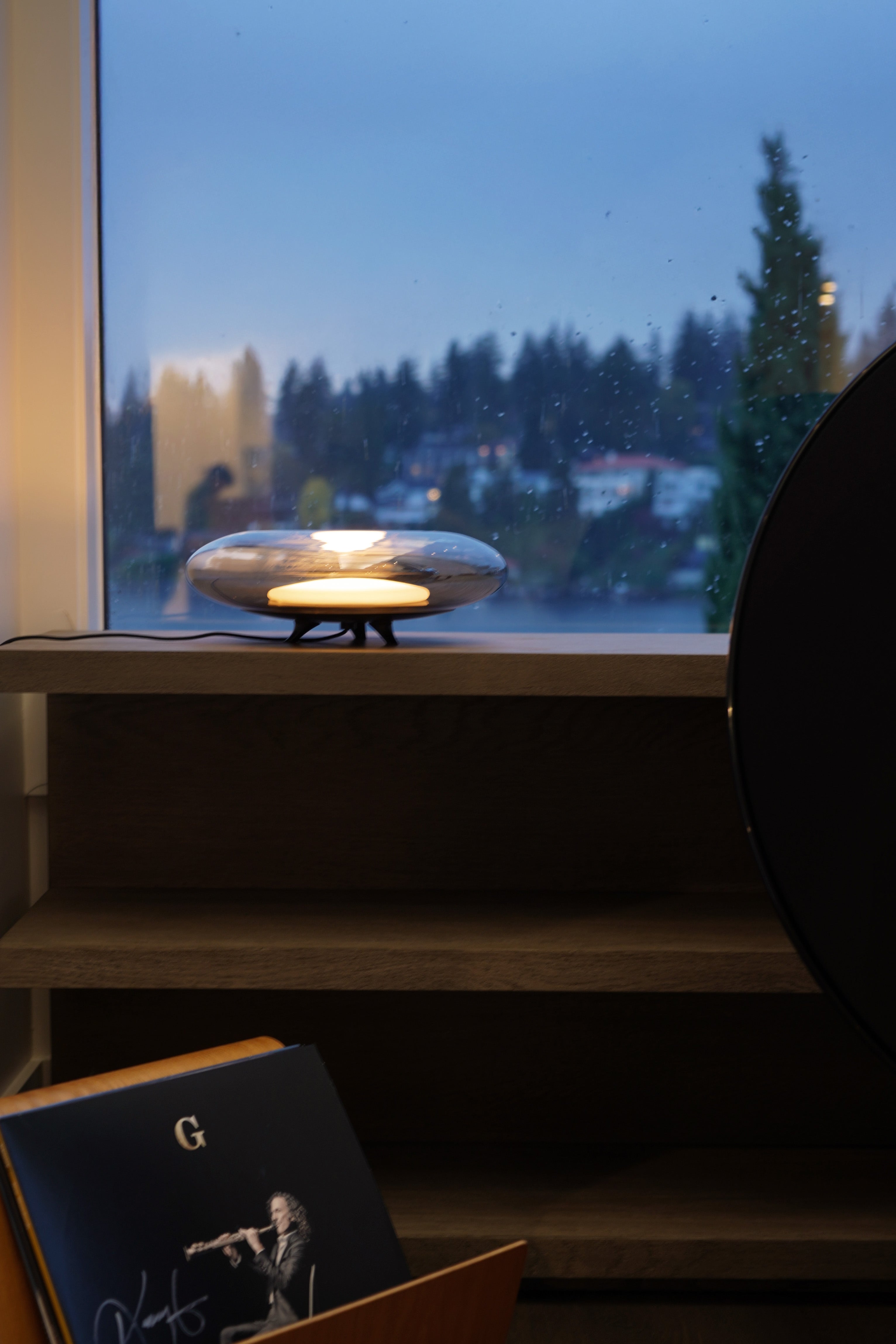 Modern lamp on a wooden surface with a window view of trees and buildings at dusk.